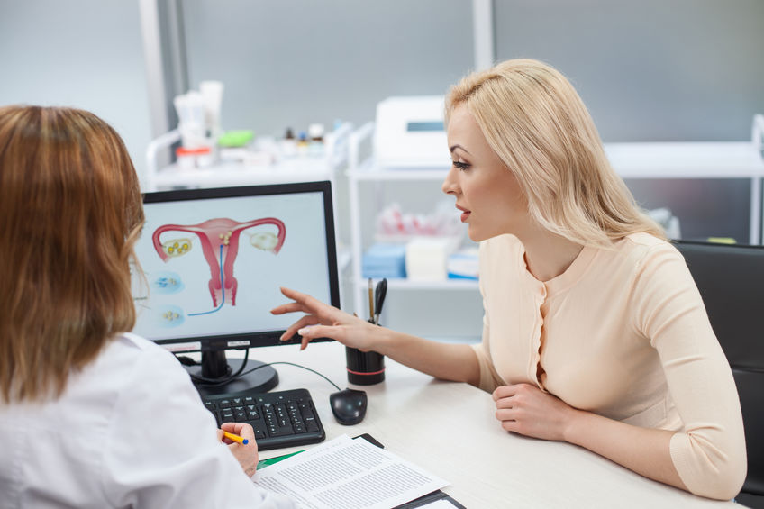 Cheerful young woman is asking advice in her gynecologist. She is sitting at the desk and pointing finger at the computer with uterus picture. Lady is listening to doctor with concentration